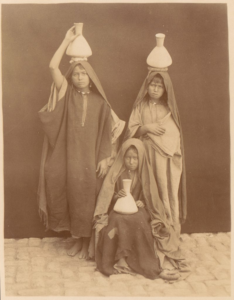 Three young Arab girls hold water jugs and pose for a photograph.